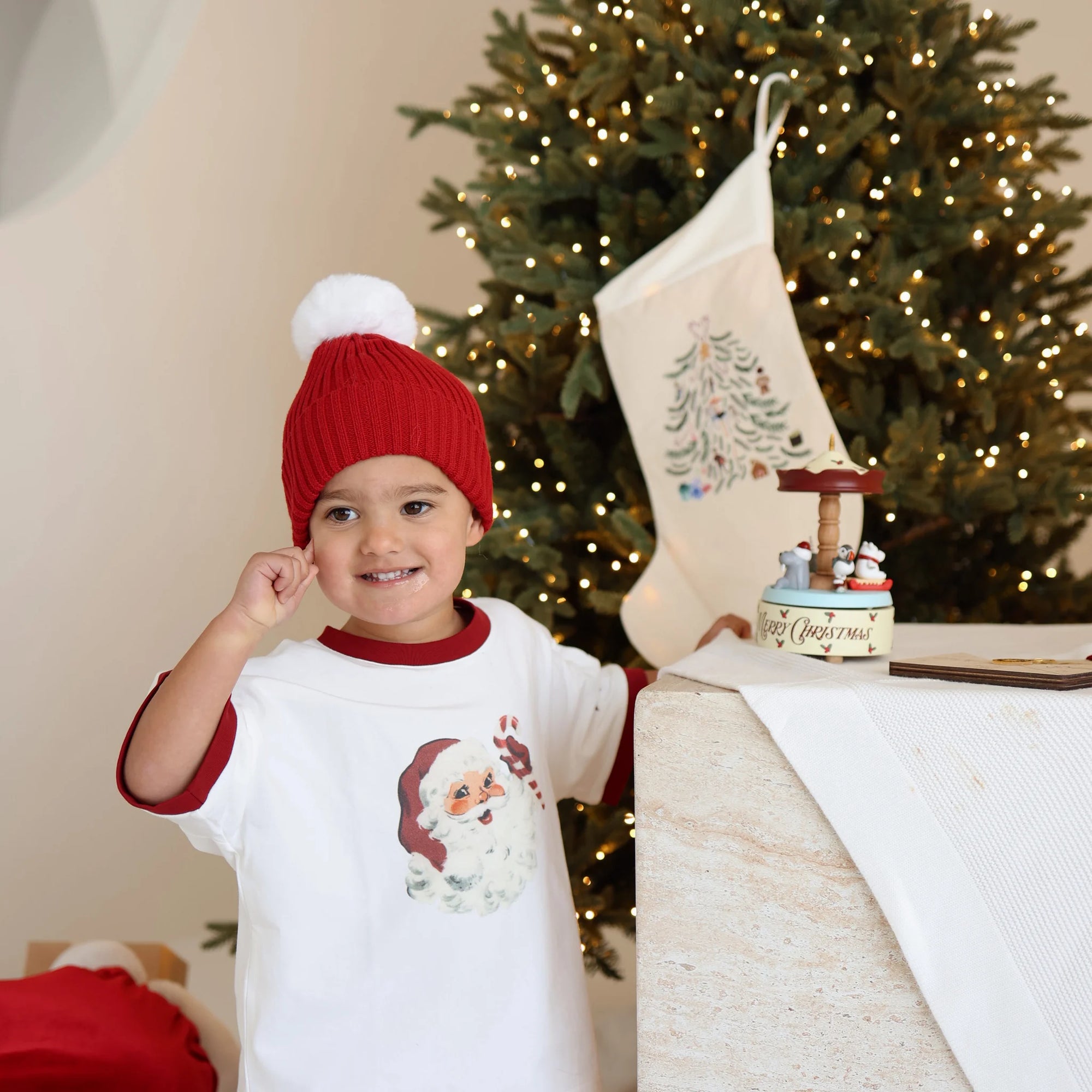 Toddler in a red pom beanie and Santa print tee smiling beside an embroidered stocking and Merry Christmas music box