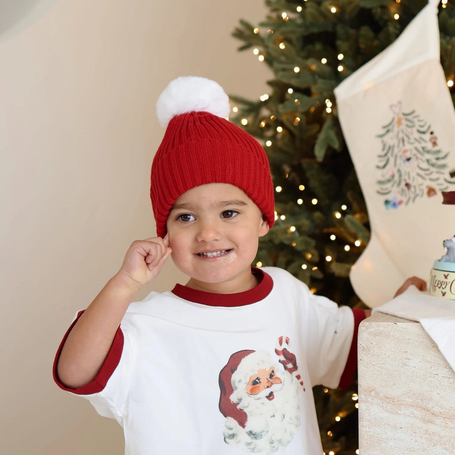 Smiling toddler in a red pom beanie and vintage Santa print tee beside an embroidered Christmas stocking