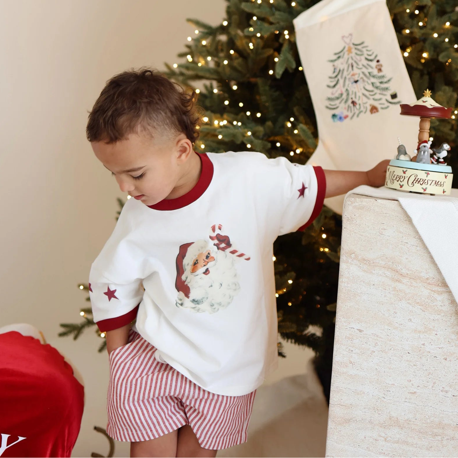 Toddler in a Santa print tee and red stripe shorts reaching for a Merry Christmas music box beside a Santa sack