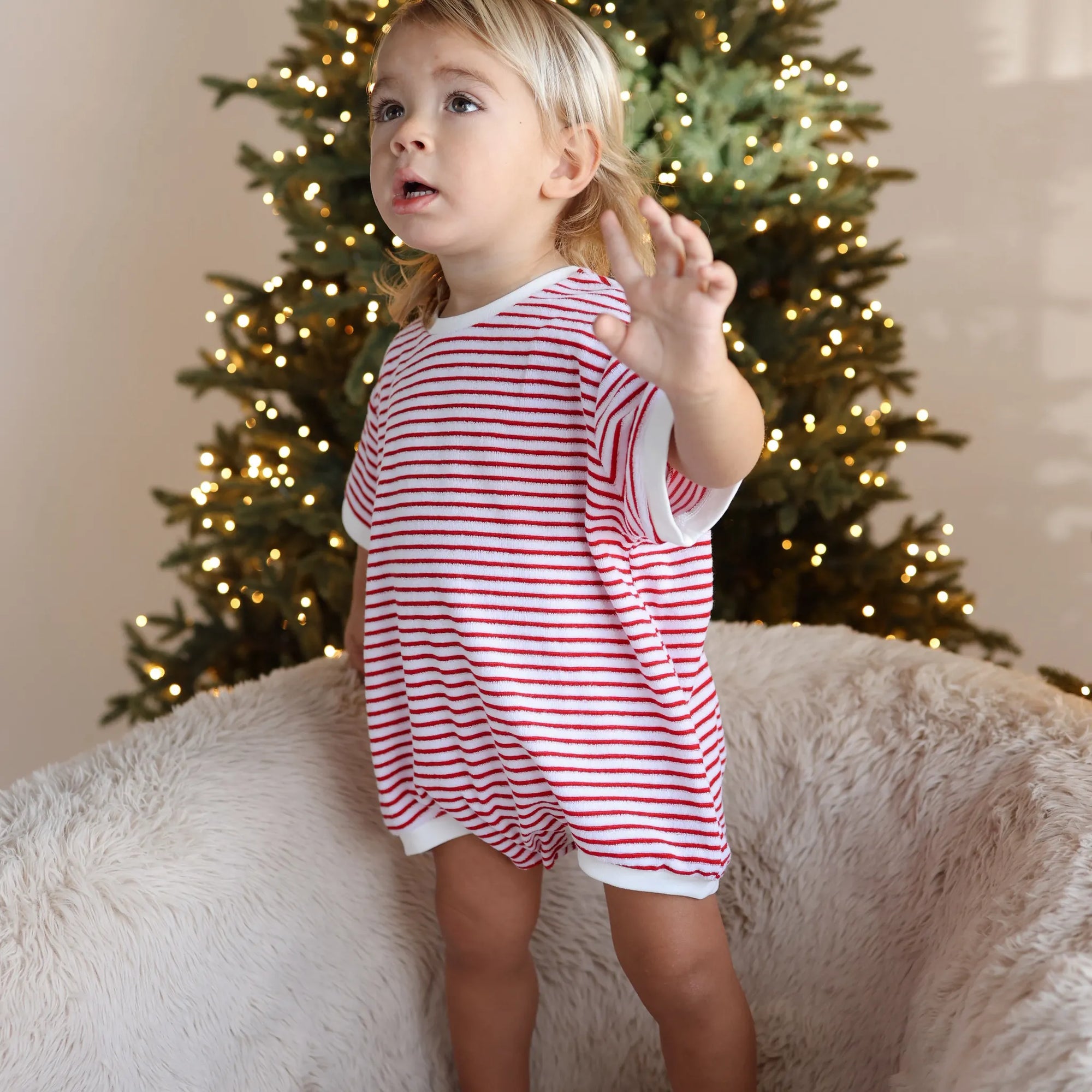 Toddler in a red and white stripe Christmas romper standing on a sheepskin chair in front of a lit Christmas tree
