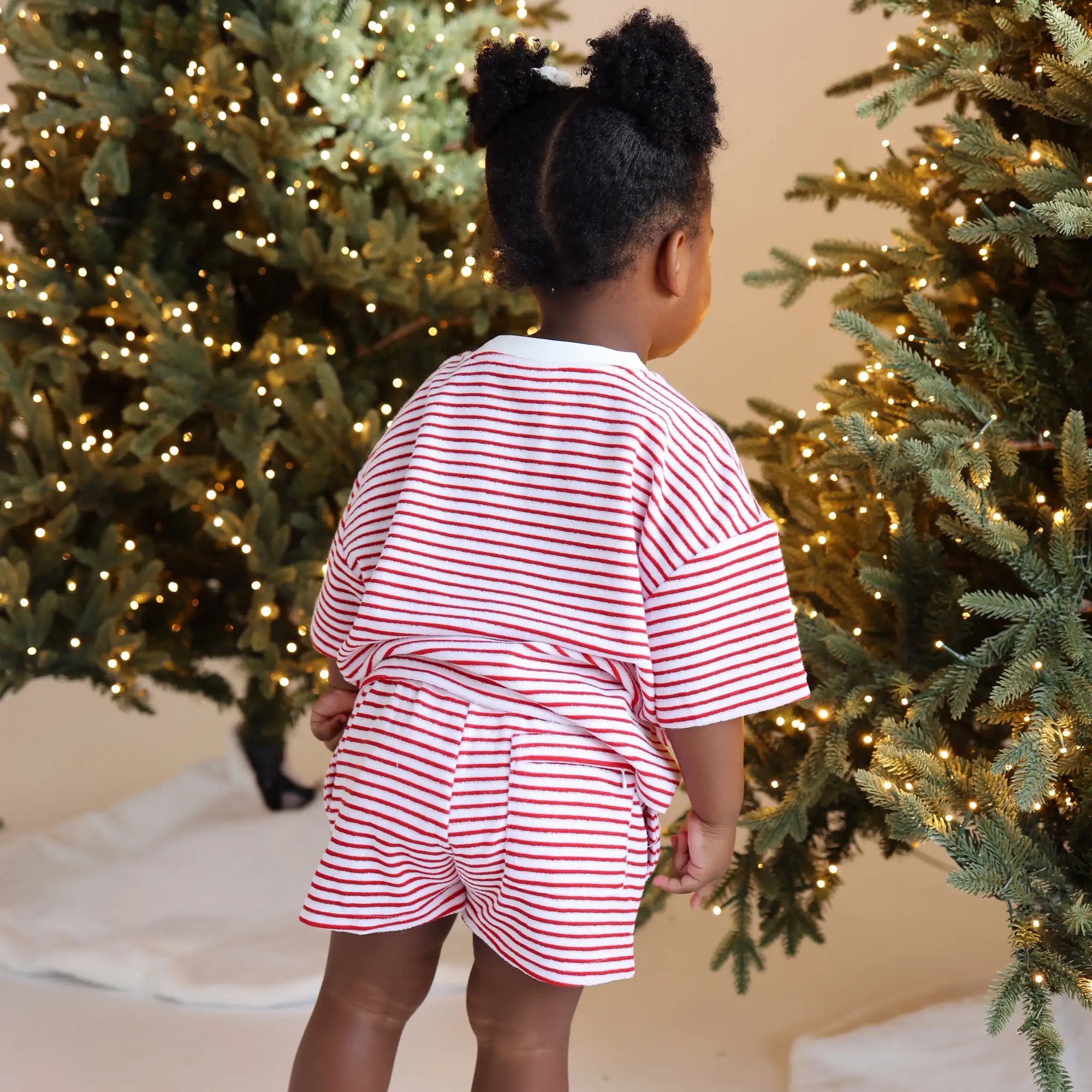Back view of a toddler in a red and white stripe Christmas pyjama tee and shorts set beside lit trees