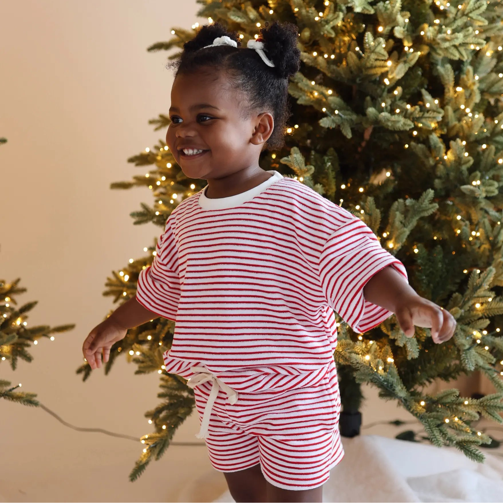 Toddler in a red and white stripe pyjama set with white hair bows, dancing in front of a lit Christmas tree
