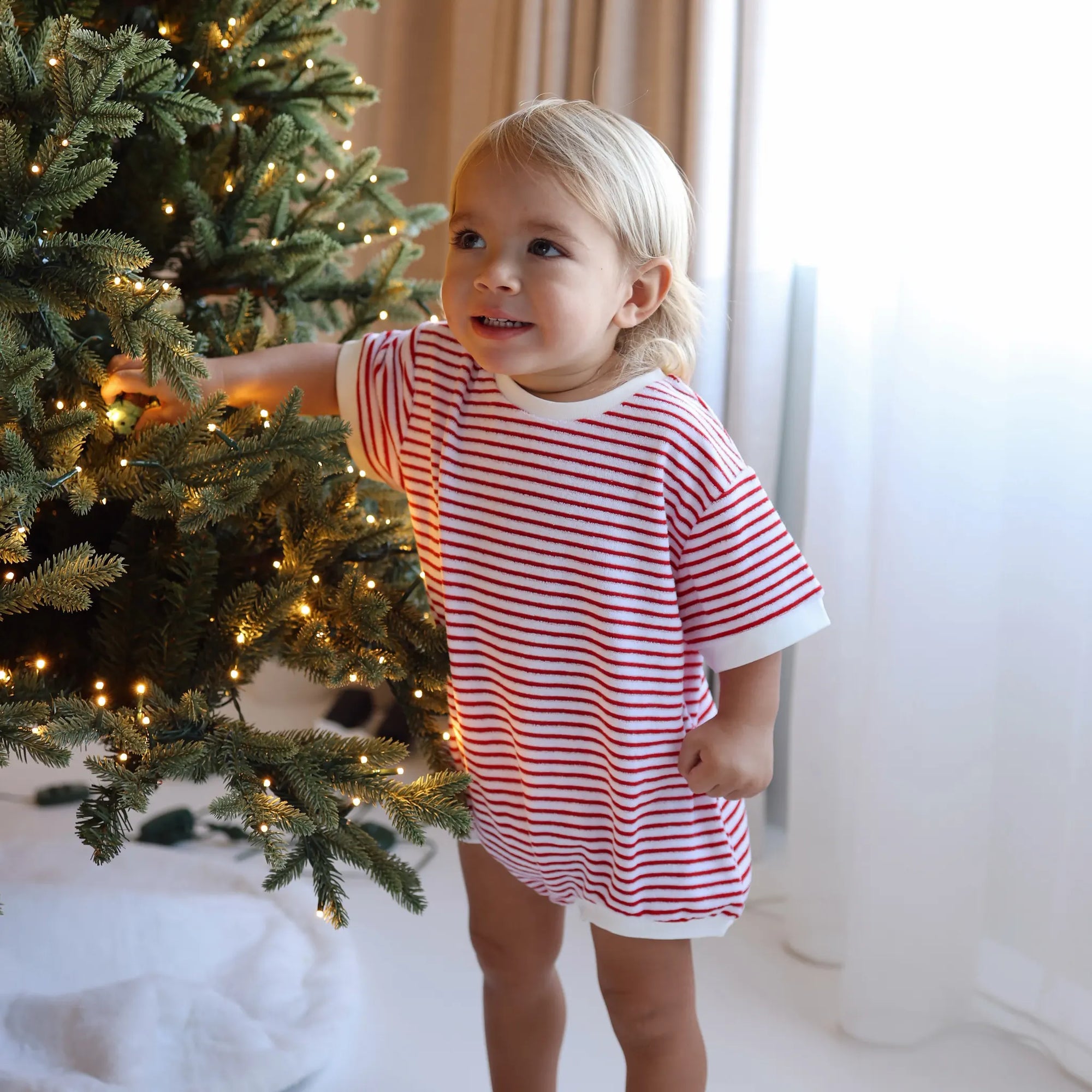 Smiling toddler in a red and white stripe Christmas romper touching the branches of a lit Christmas tree
