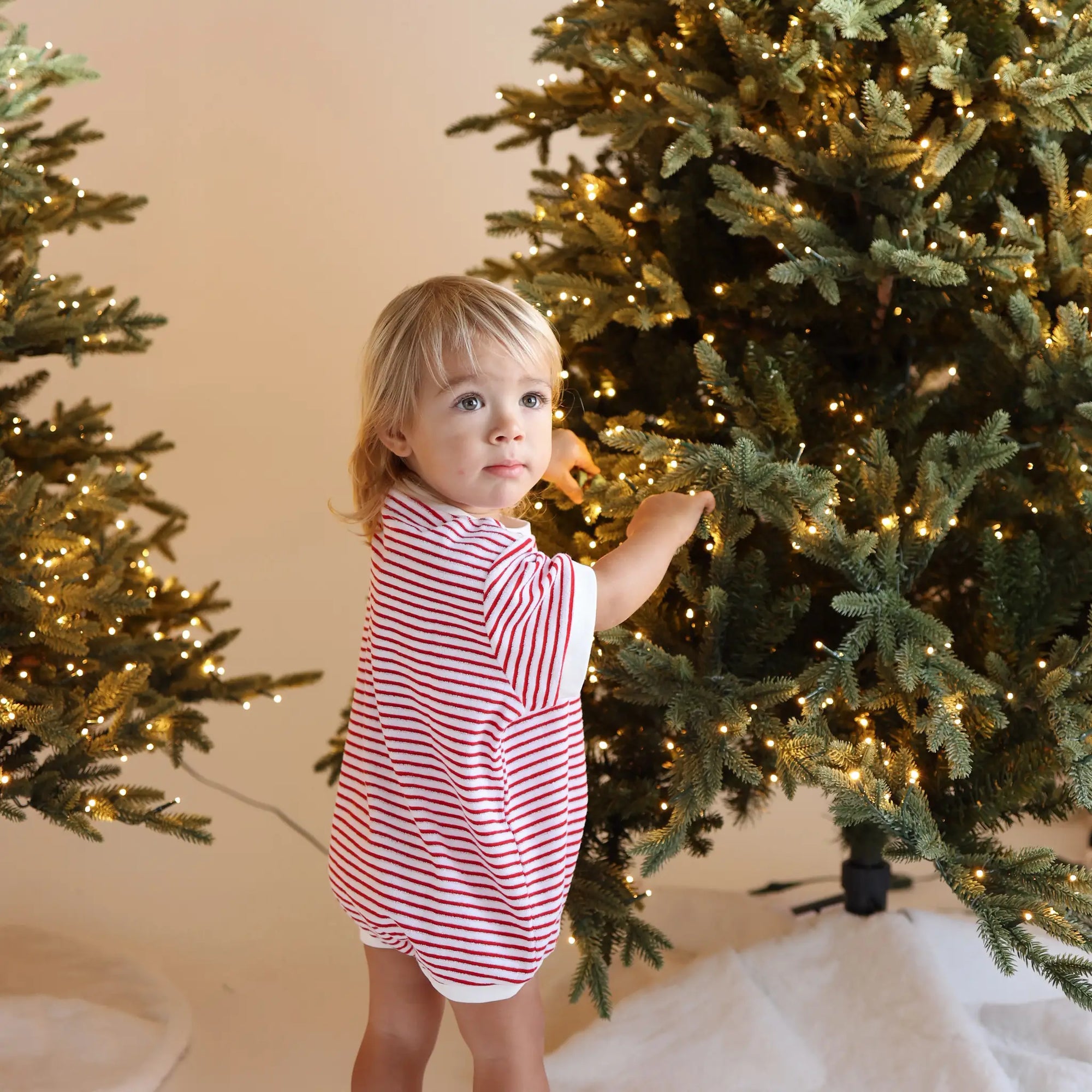 Toddler in a red and white stripe Christmas romper looking over her shoulder beside two lit Christmas trees