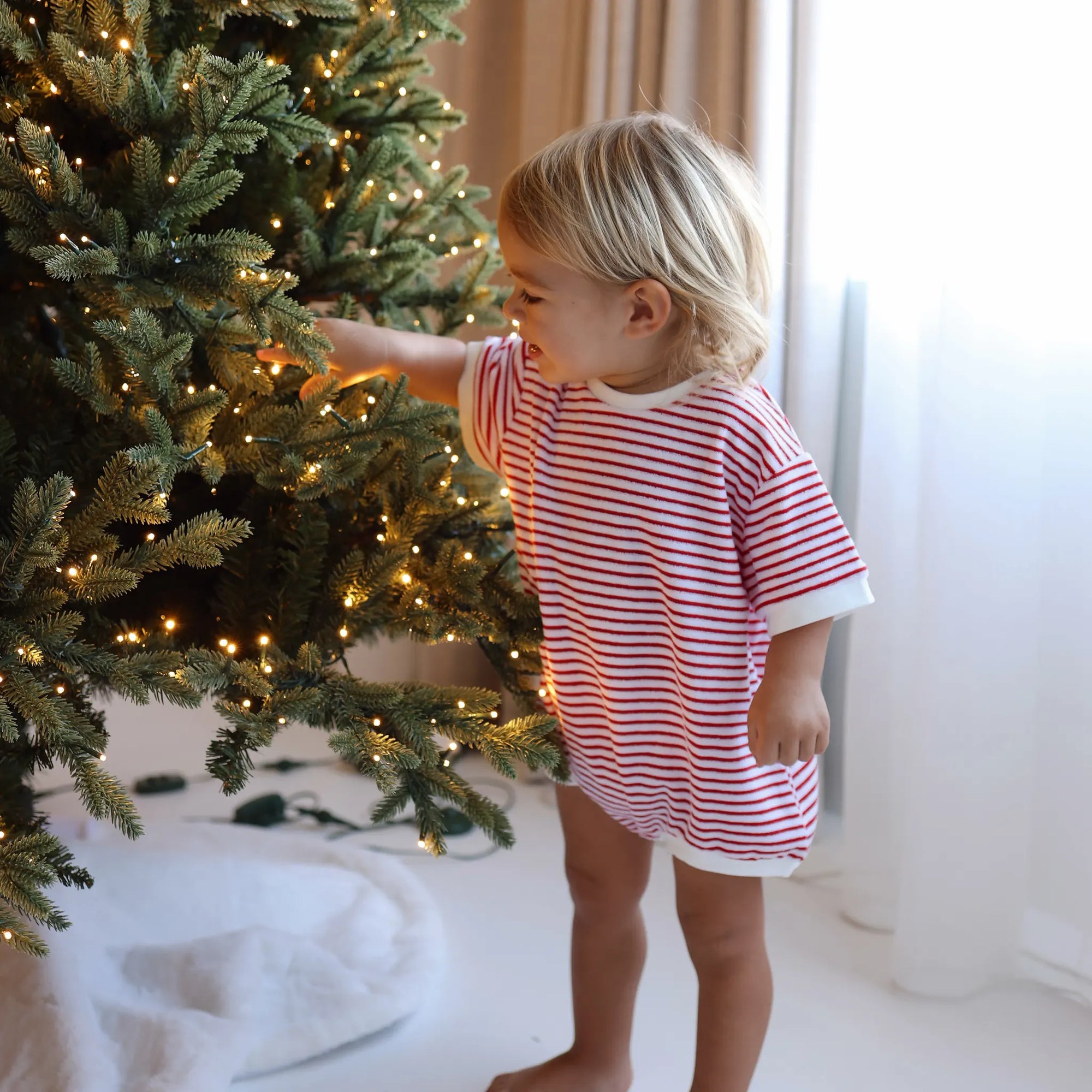 Toddler in a red and white stripe Christmas romper reaching up to decorate a lit Christmas tree