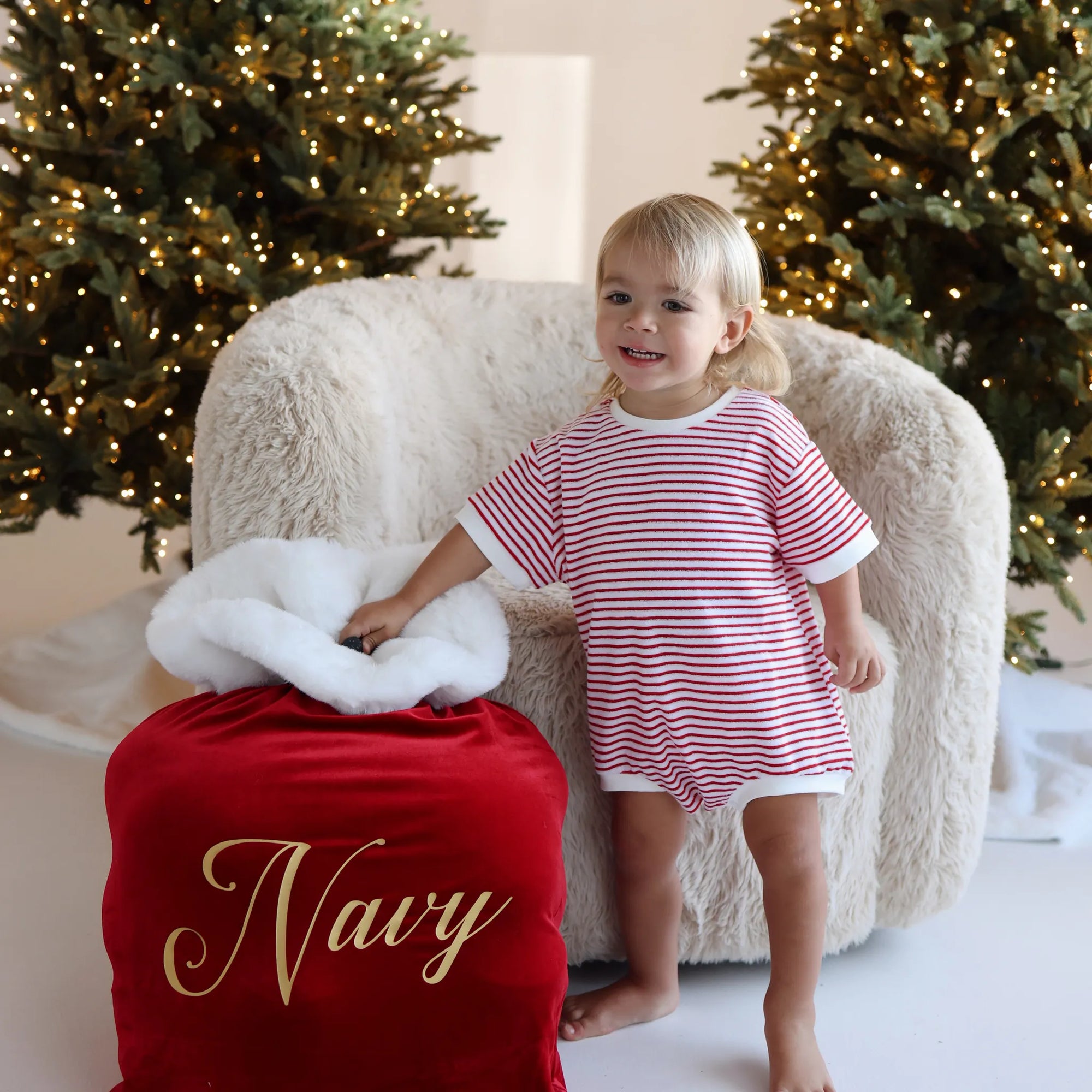 Toddler in a red and white stripe Christmas romper beside a personalised red velvet Santa sack embroidered with the name Navy