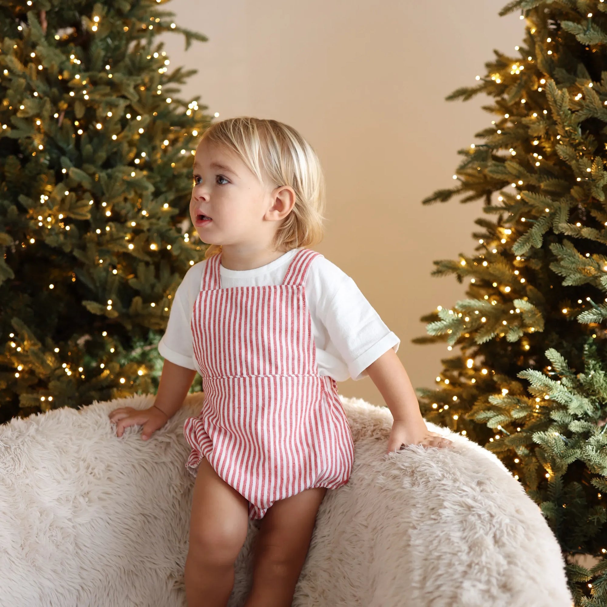 Toddler in a red stripe overall romper and white tee kneeling on a sheepskin chair between lit trees