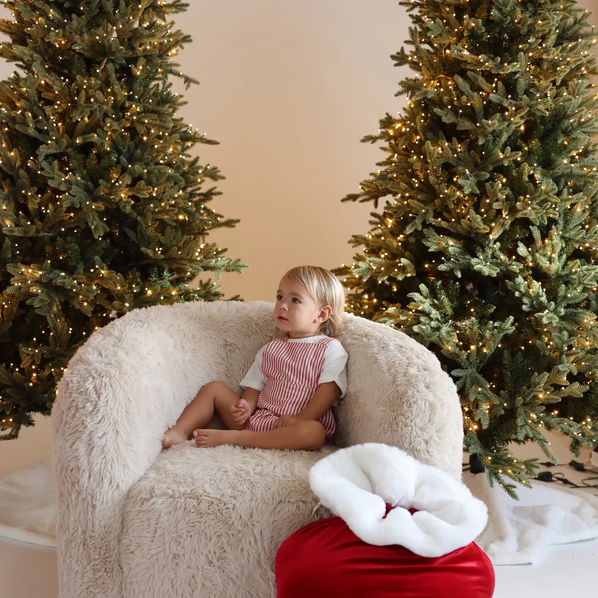 Toddler in a red stripe overall romper sitting on a sheepskin chair beside a red Santa sack