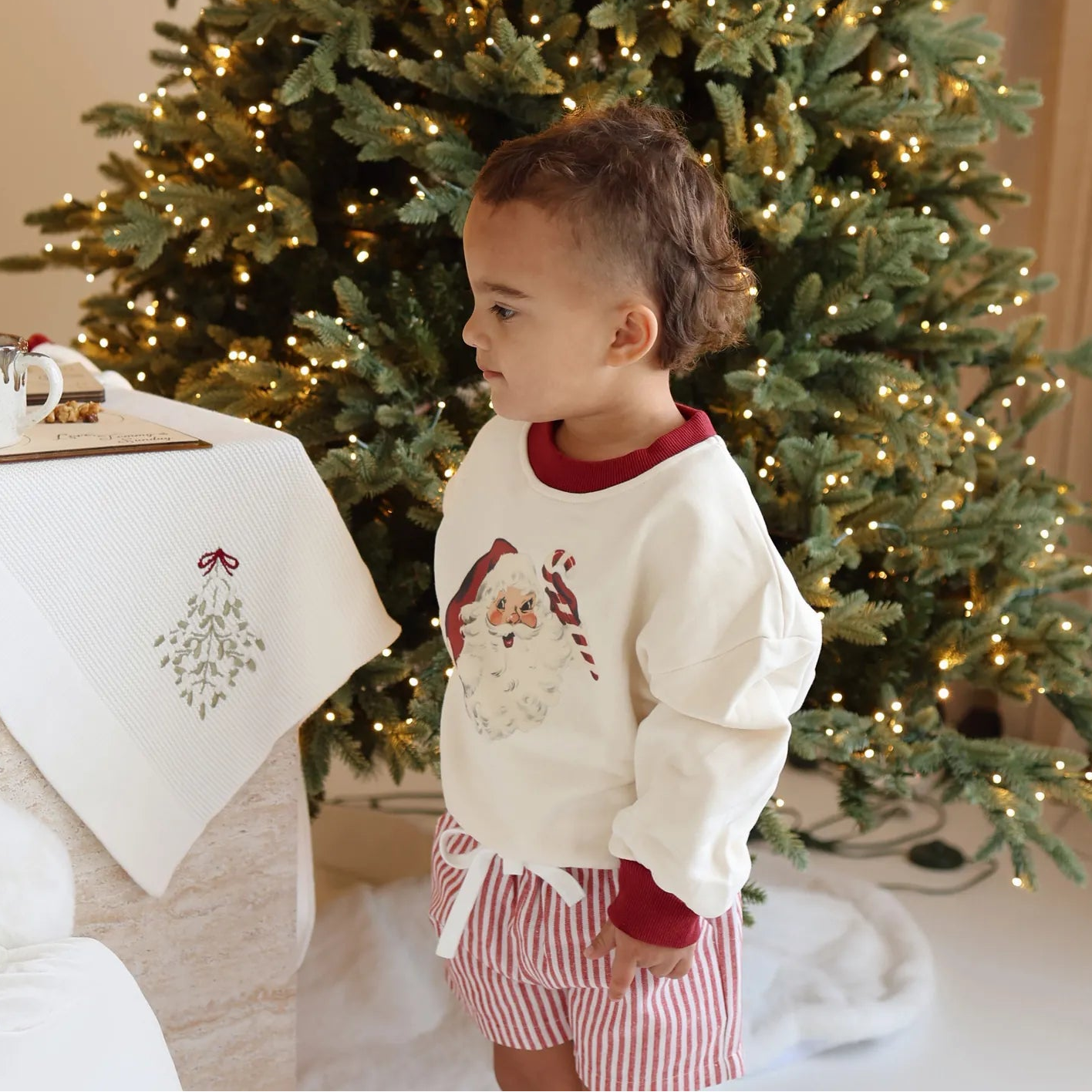 Toddler in a vintage Santa print sweatshirt and red stripe shorts standing beside a table set with milk and cookies