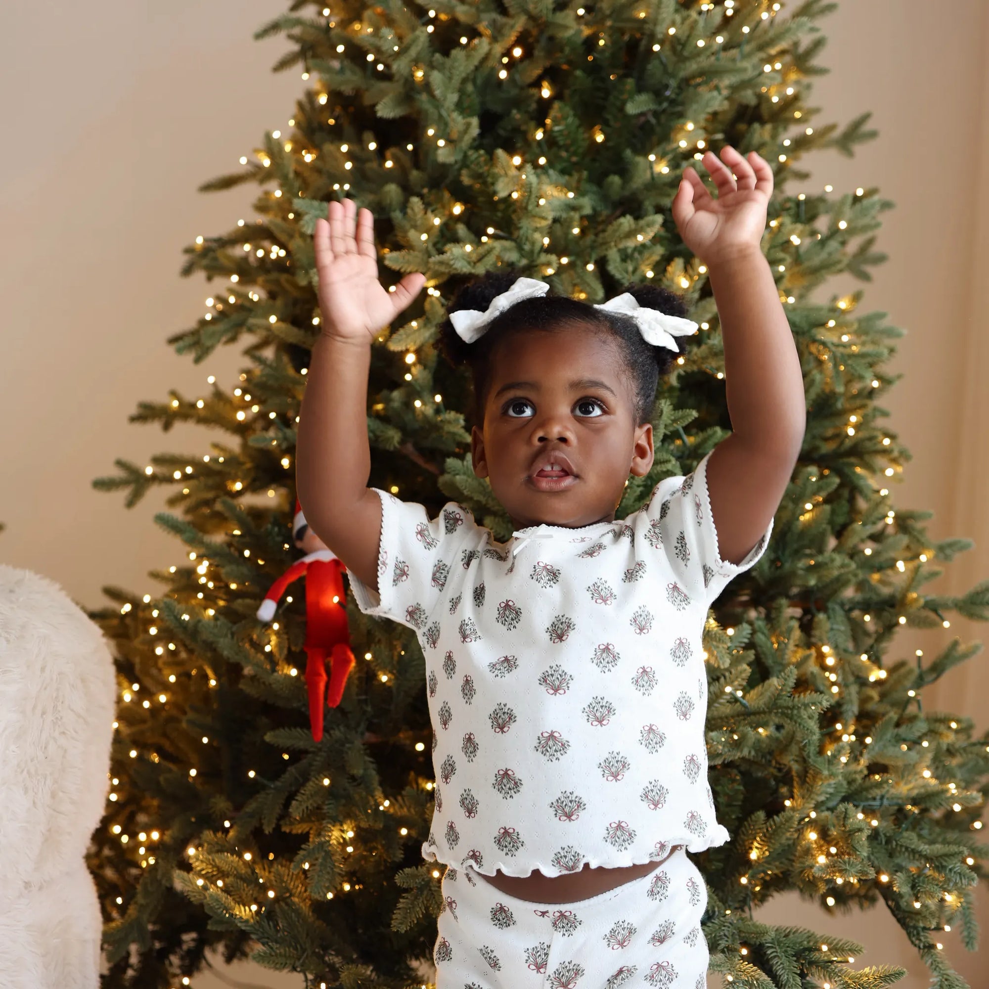 Toddler in wreath print pyjamas with white hair bows reaching both arms up in front of a lit Christmas tree