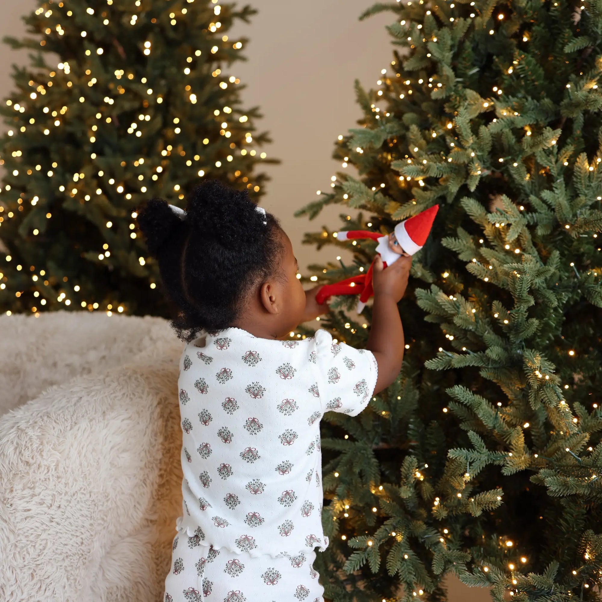 Toddler in wreath print pyjamas placing an elf doll into a lit Christmas tree