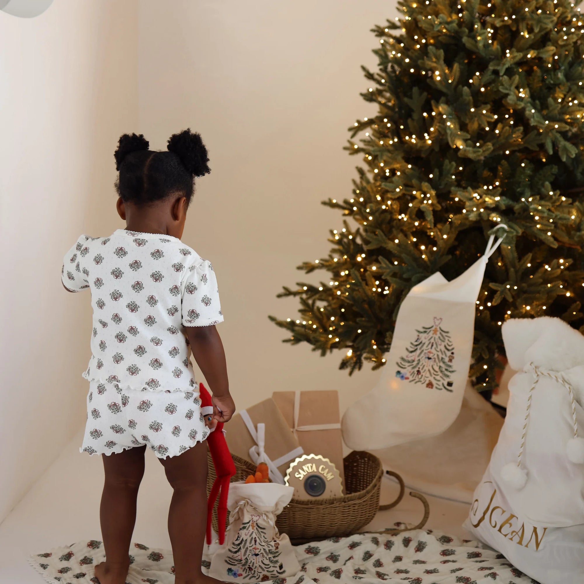 Toddler in wreath print pyjamas holding an elf doll beside embroidered stockings and a personalised Santa sack