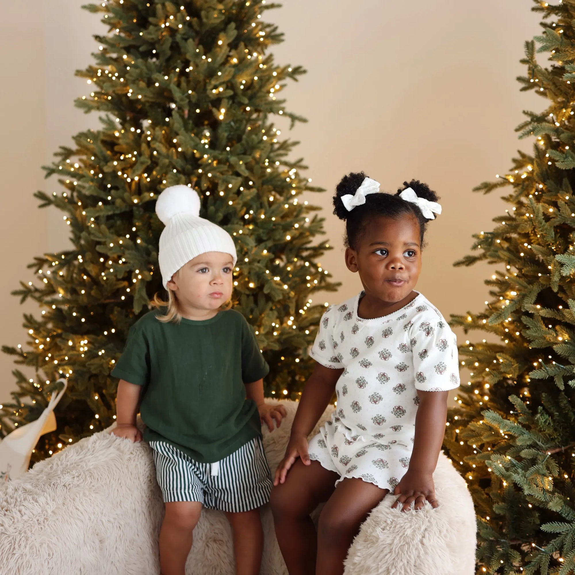 Two toddlers between lit Christmas trees, one in a green tee and stripe shorts with a white pom beanie, one in a wreath print set with white hair bows
