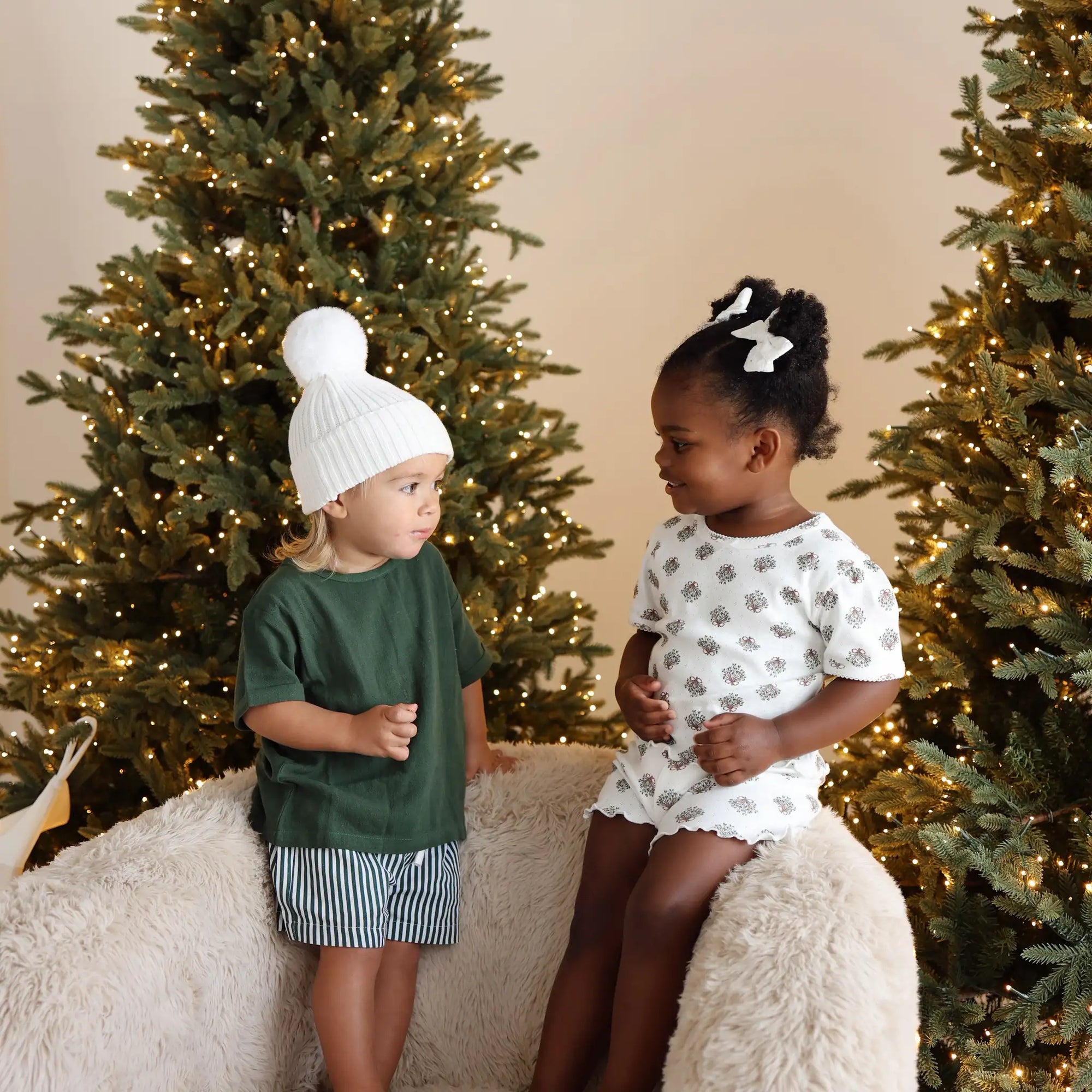 Two toddlers between lit Christmas trees, one in a green tee and stripe shorts with a white pom beanie, one in wreath print pyjamas