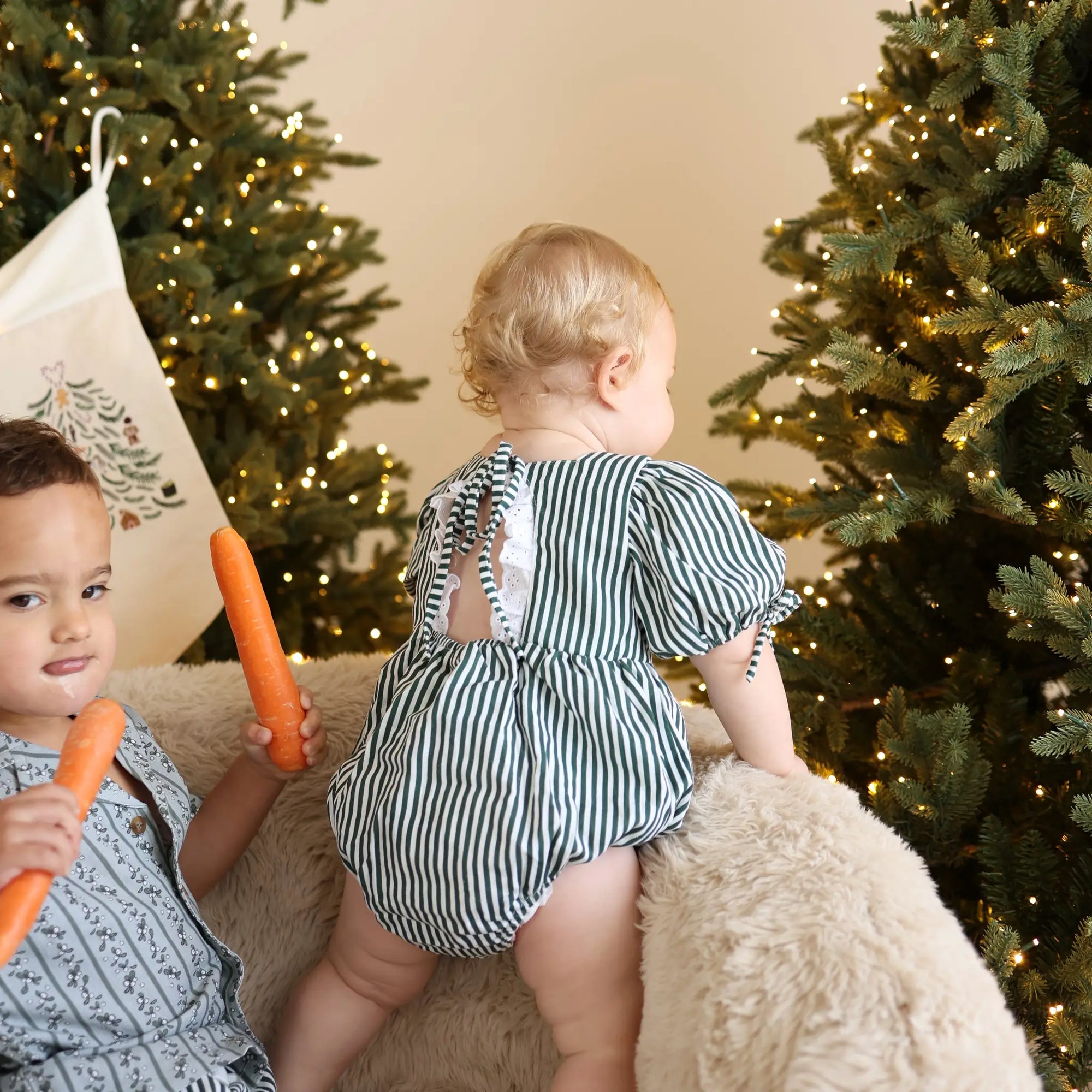 Back view of a baby in a green stripe romper with tie back detail beside a toddler holding carrots