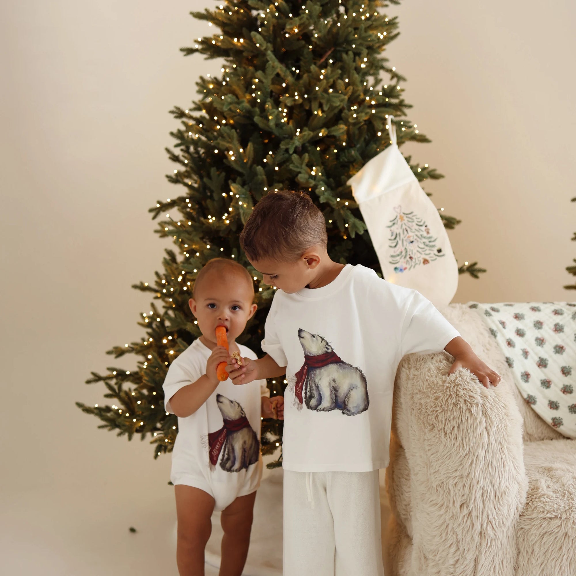 Two children in polar bear print pieces, a baby in a romper and a boy in a tee and cream pants, by a lit tree