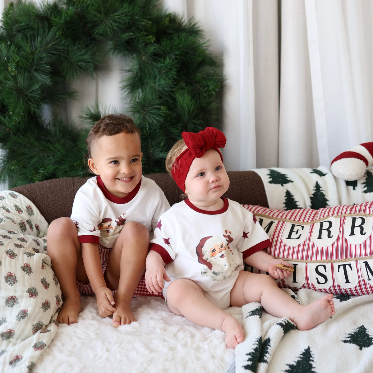 Two toddlers in vintage Santa print tee and short sets sitting on a sofa with a Christmas wreath and Merry Christmas cushions