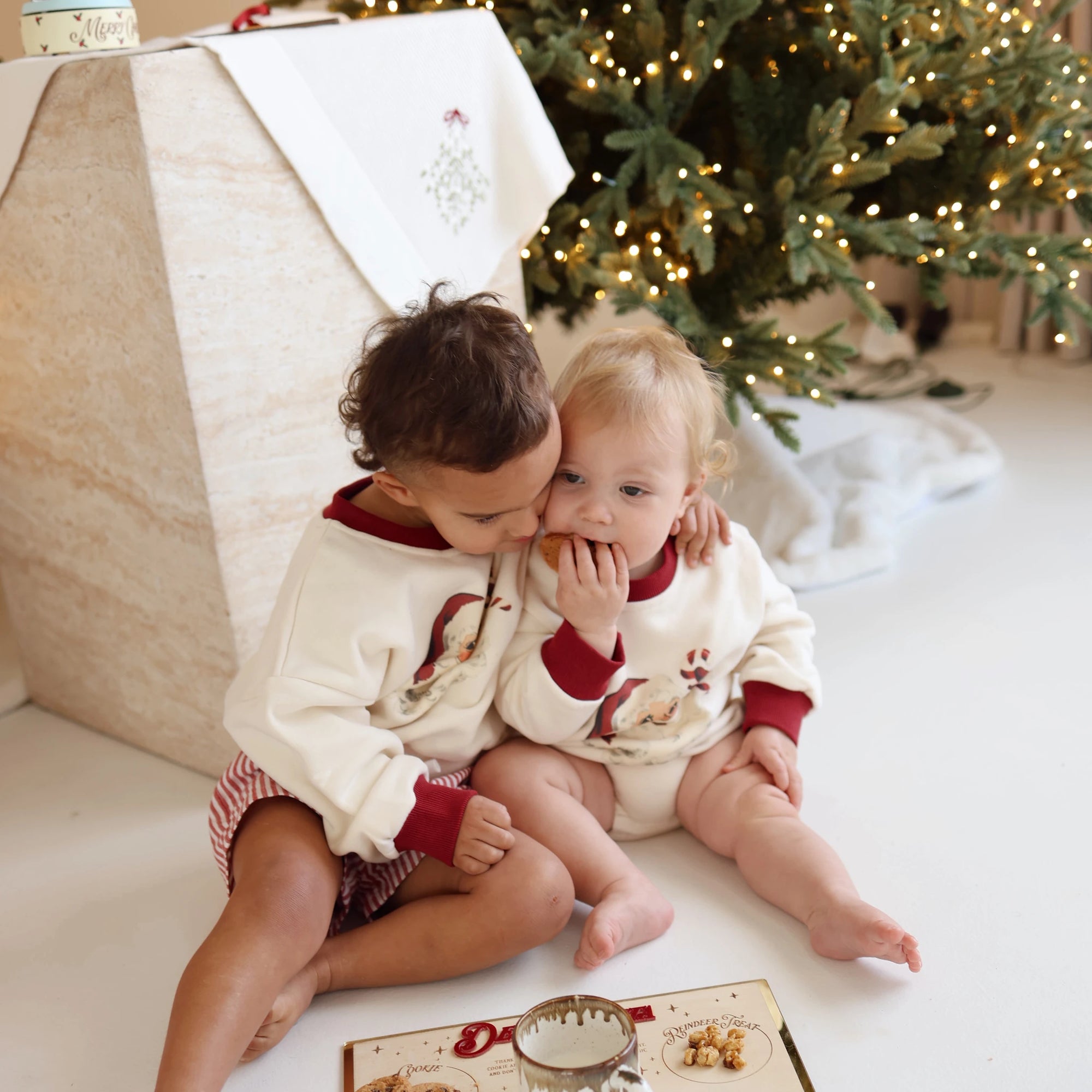 Toddler kissing a baby on the cheek, both in vintage Santa print jumpers, beside a lit Christmas tree