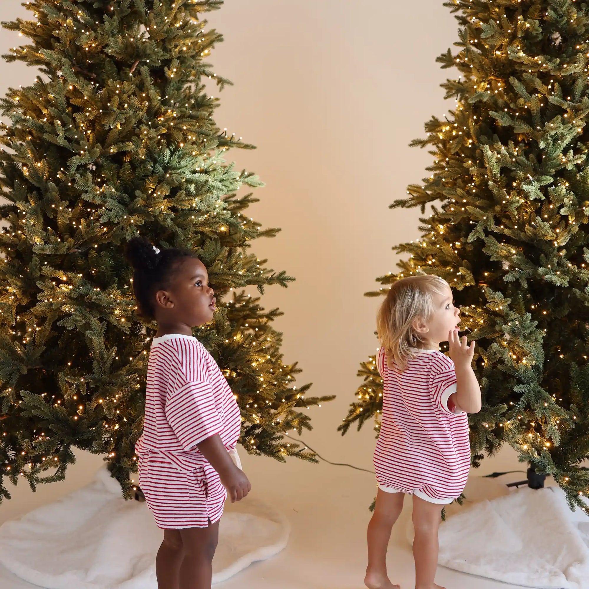 Two toddlers in red and white stripe Christmas pyjamas looking up at lit Christmas trees