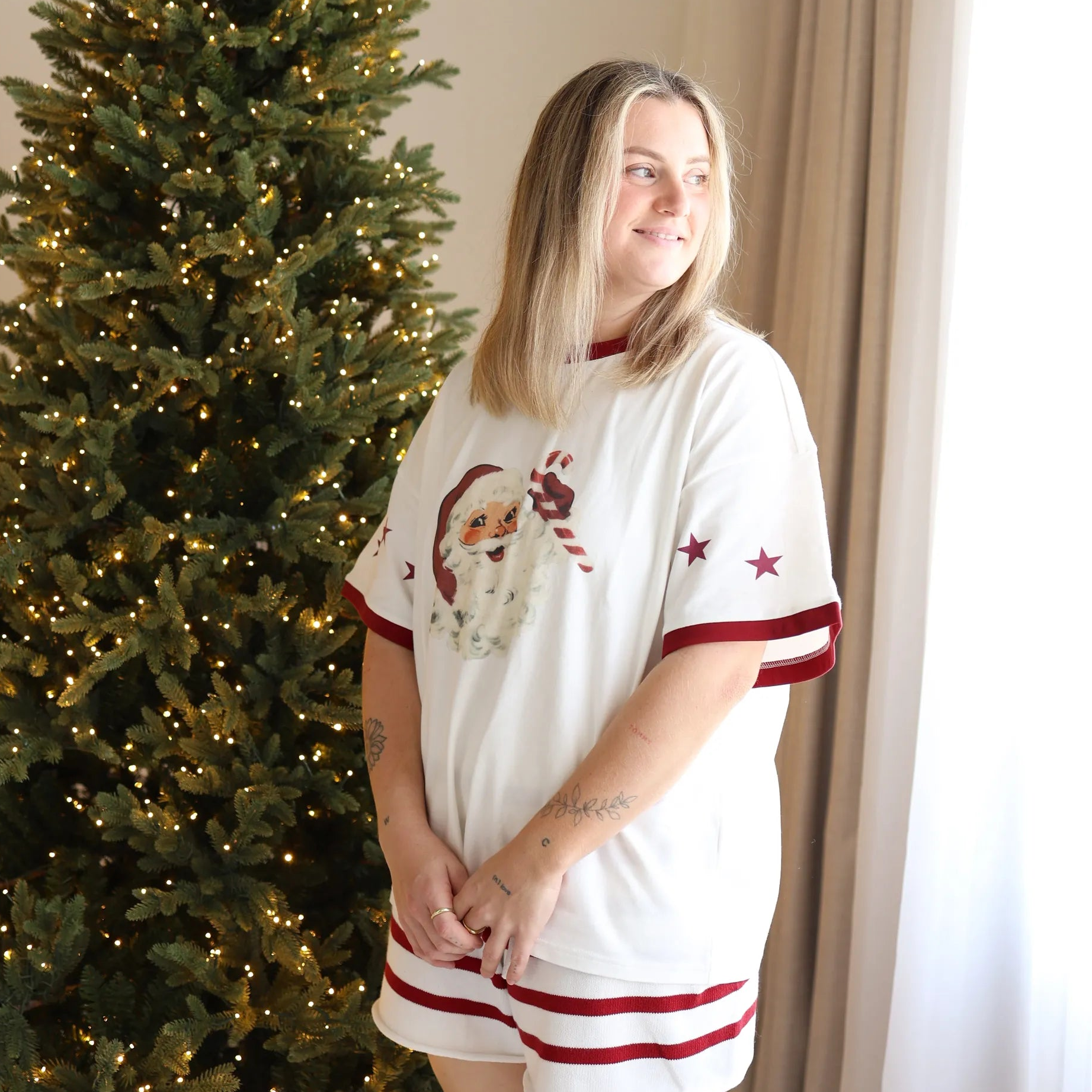 Woman in a vintage Santa print tee and red stripe shorts pyjama set beside a lit Christmas tree