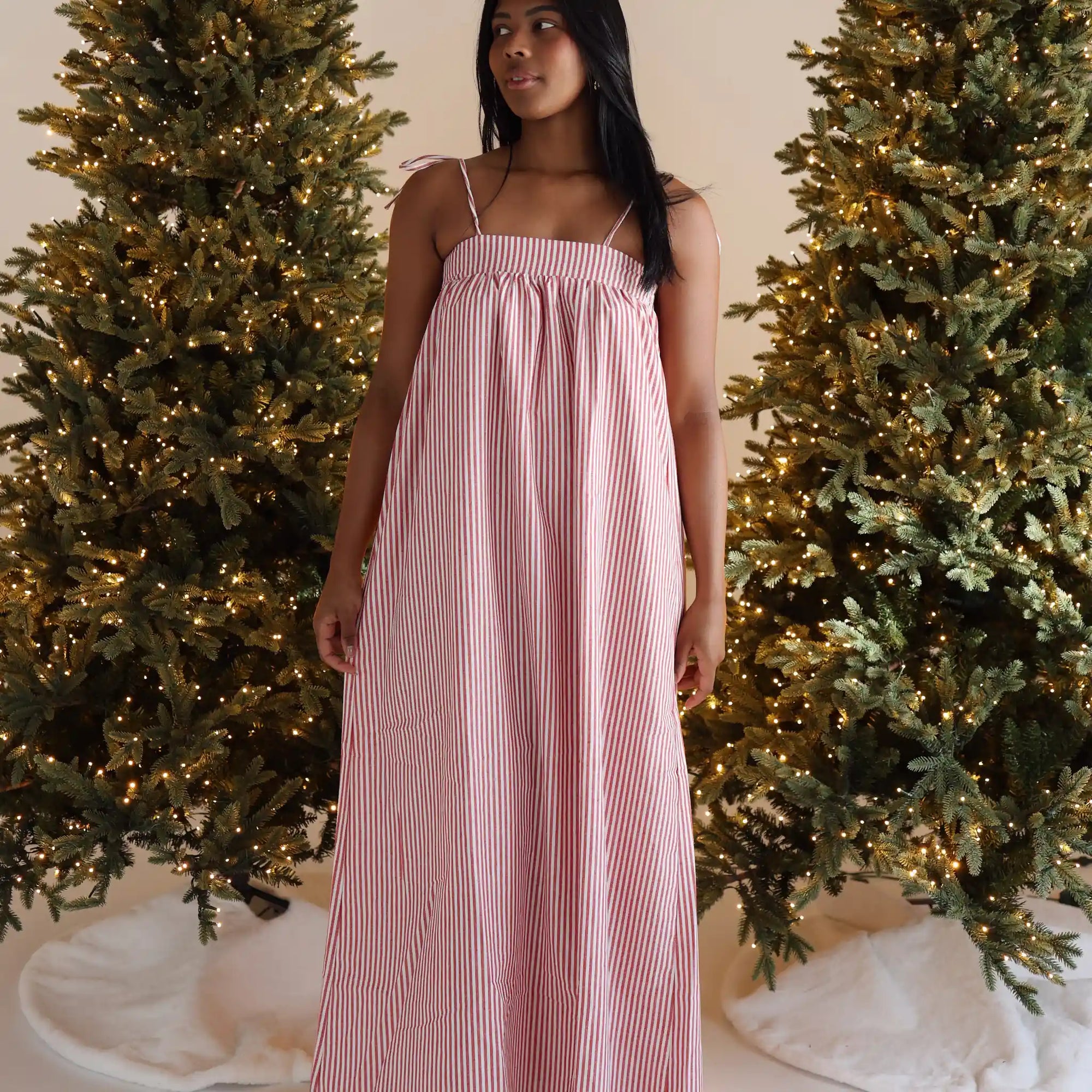 Woman in a red and white stripe shirred maxi Christmas dress between two lit Christmas trees